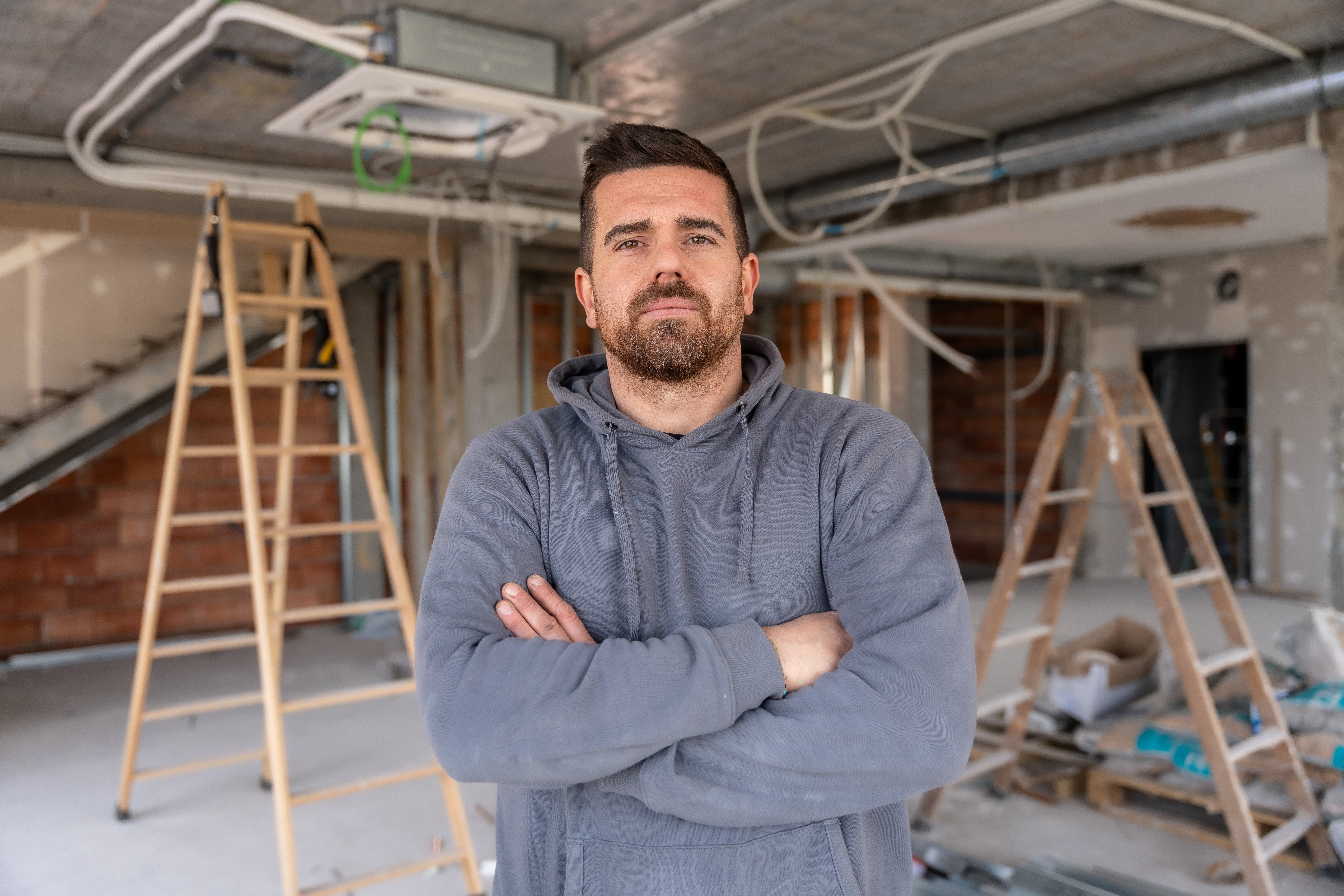 Contractor standing with arms crossed in a construction site, looking at the camera while showing the progress of an air conditioning installation in the ceiling